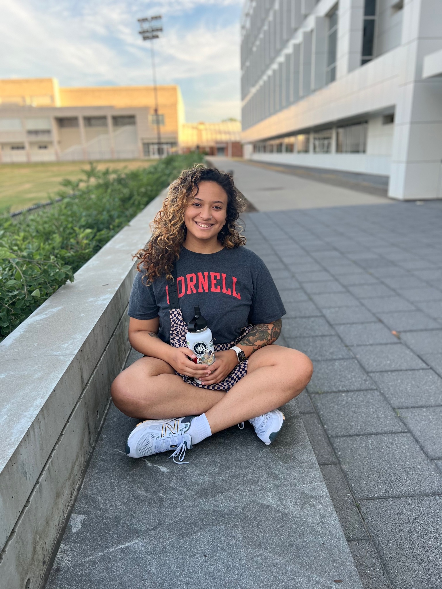 Aalayna, Cornell shirt, sitting cross legged on the ground, outside, smiling