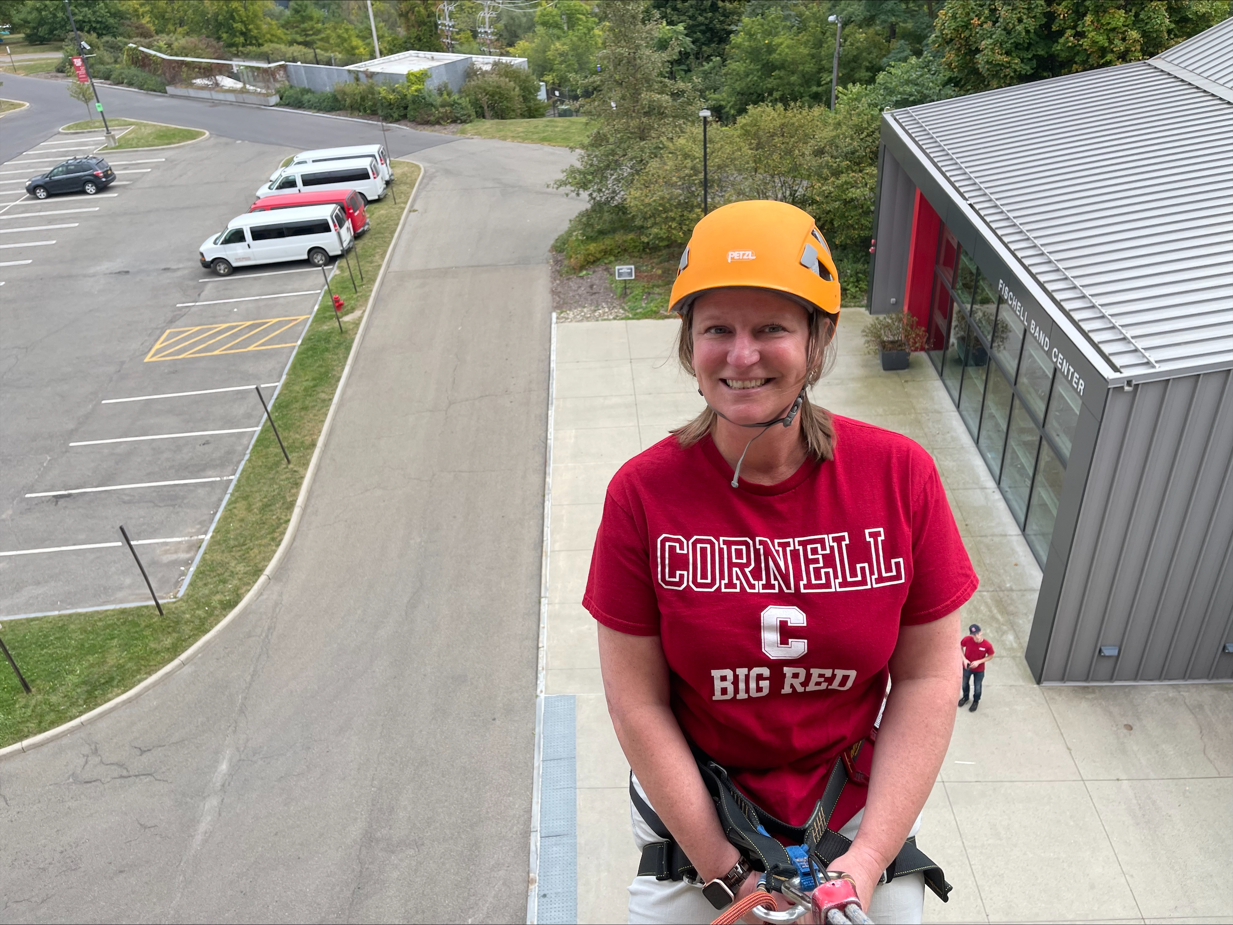 Amy in a red shirt and yellow helmet and rock climbing gear smiling up at the camera 